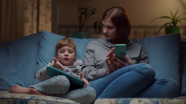 30s femme et jeune garçon regardant tablette et téléphone portable assis sur un canapé confortable le soir. Enfant jouer et maman surfer en ligne sur des gadgets à la maison . — Video