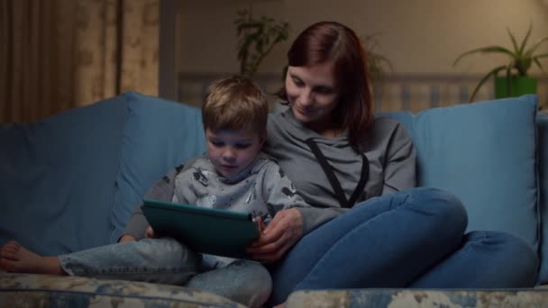 30s femme et jeune garçon regardant ensemble tablette d'ordinateur assis sur un canapé confortable le soir. Enfant et maman regardant en ligne sur gadget à la maison . — Video