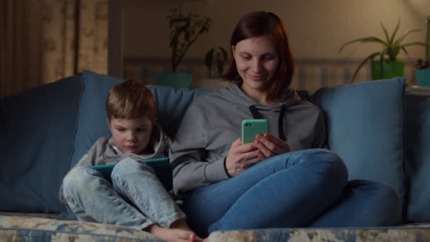 Mujer de 30 años y niño joven viendo tableta y teléfono móvil sentado en un sofá acogedor por la noche. Niño jugando y mamá surfeando en línea en gadgets en casa . — Vídeos de Stock