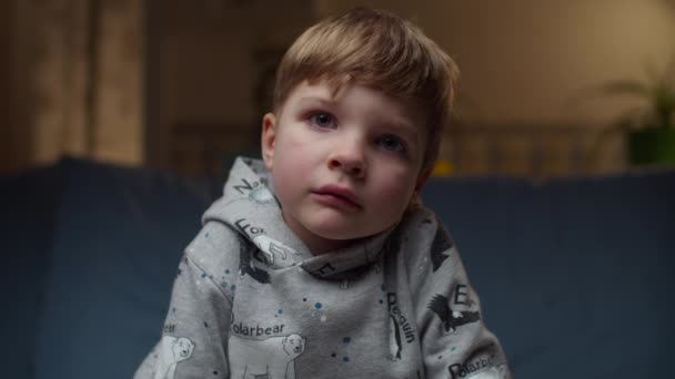 Porträtt av en ung upprörd pojke som sitter på soffan hemma. Tråkiga barn ansikte tittar på kameran i slow motion. — Stockvideo