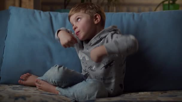 ブロンドの男の子が夜の部屋で居心地の良いソファの上に座ってバカ。1人でリビングで遊んでいるジーンズの子供。面白い子作るダンスムーブメントオンソファ. — ストック動画