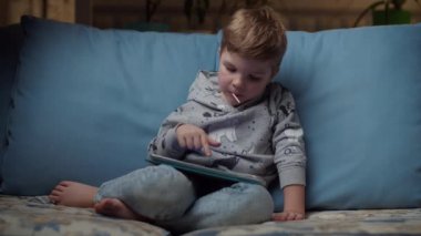 Ağzında lolipopu olan genç bir çocuk gece rahat bir koltukta oturup tablet bilgisayar izliyor. Cihazla oynayan çocuk. 