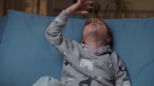 Jonge jongen neemt lolly naar de mond zitten op gezellige bank 's avonds. Kind genieten van hard snoep en glimlachen in slow motion. — Stockvideo