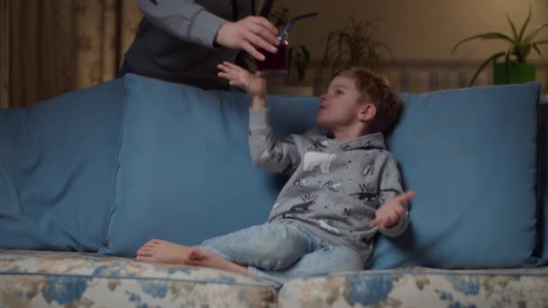 Blond preschooler boy drinking juice sitting on cozy couch at evening. Mother brings son glass of drink with straw. Kid drinking blueberry juice with non-plastic straw on sofa at home. — Stock Video