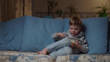 Sarışın anaokulu çocuğu akşam rahat bir kanepede öğle yemeği yiyor. Çocuk kaşıkla beyaz pirinç yemekten hoşlanıyor. Kotlu çocuk TV karşısında kanepede. 