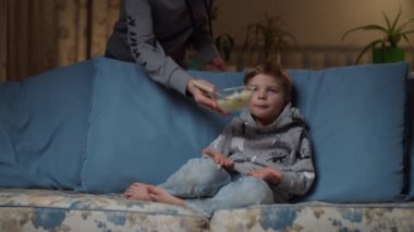 Sarışın anaokulu öğrencisi akşam rahat koltukta oturuyor ve annesi ona bir kase yemek getiriyor. Çocuk kaşıkla beyaz pirinç yiyor. Anneler, oğullarına tabak veriyor..