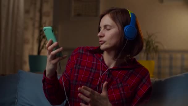 Молода 30 років жінка в червоній сорочці слухає музику в кольорових навушниках, сидячи на дивані вдома. Жінка насолоджується музикою із закритими очима у повільному русі . — стокове відео