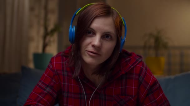 Νεαρή 30άρα γυναίκα με κόκκινο πουκάμισο σε χρωματιστά ακουστικά που μιλάει στην κάμερα που κάθεται στον καναπέ στο σπίτι. Γυναικείες ιστορίες που κοιτάζουν την κάμερα σε αργή κίνηση. — Αρχείο Βίντεο
