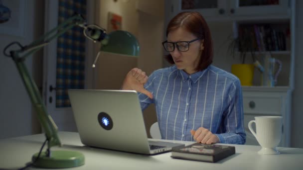 30 'lu yaşlardaki gözlüklü genç bayan öğretmen başparmağıyla evde dizüstü bilgisayara bakarak duygusal konuşuyor. Çevrimiçi eğitim süreci. Evdeki insanlar çalışıyor.. — Stok video