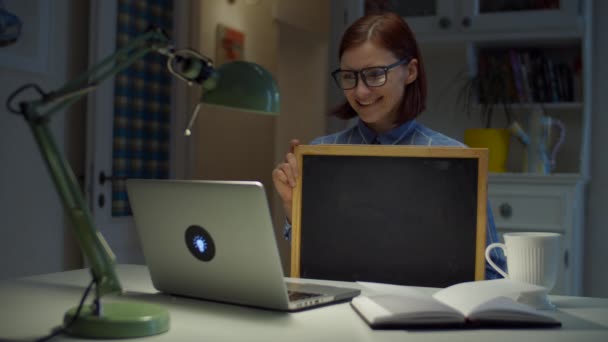 Jovem mulher de 30 anos de óculos segurando limpo placa de giz vazio olhando para laptop em casa. Processo de educação online. Trabalho a partir de casa pessoas . — Vídeo de Stock