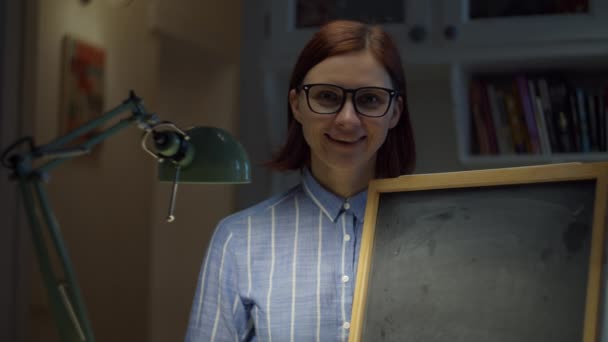 30s woman in glasses holding empty chalk board looking at camera at home. Online education process. Webcam view of teacher talking. — Stock Video