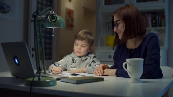 Giovane madre degli anni '30 che fa i compiti online con il figlio in età prescolare a casa. Donna in occhiali e ragazzo che studia online con laptop, notebook e libro di testo. Processo educativo online . — Video Stock