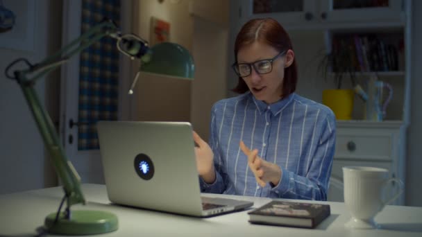Молодая 30-летняя учительница в очках разговаривает и жестикулирует эмоционально глядя на ноутбук дома. Процесс онлайн-обучения. Работа на дому — стоковое видео
