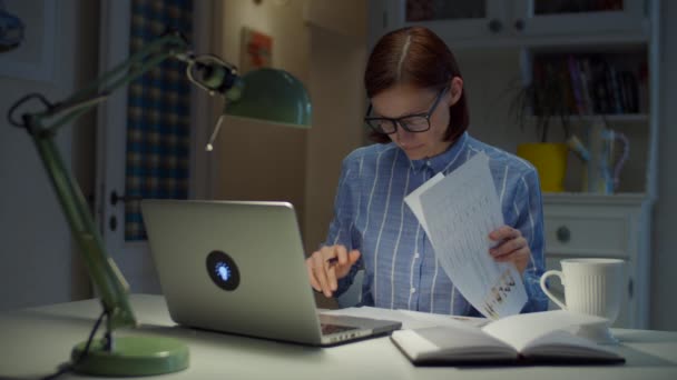 Νεαρή 30χρονη που εργάζεται από το σπίτι γυναίκα κοιτάζει μέσα από έγγραφα και πινακίδες με στυλό. Διαδικτυακή εκπαίδευση. Επιχειρηματίας σε γυαλιά με φορητό υπολογιστή στο σπίτι. — Αρχείο Βίντεο