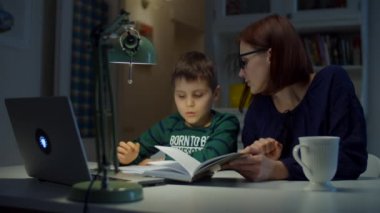 30 'lu yaşlarda genç bir anne, okullu oğluyla çevrimiçi ödev yapıyor. Gözlüklü bir kadın ve dizüstü bilgisayarı, defteri ve ders kitabı olan bir çocuk. Çevrimiçi eğitim süreci. 