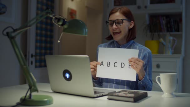 Молодая 30-летняя учительница в очках разговаривает и показывает лист бумаги с буквами алфавита, глядя на ноутбук дома. Процесс онлайн-обучения. Работа на дому . — стоковое видео