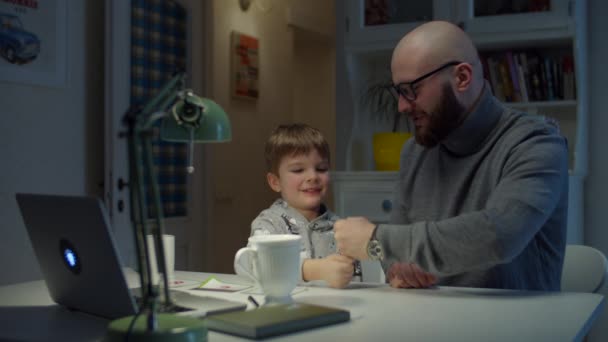 Giovane padre anni '30 dà cinque al figlio, mentre facendo i compiti online a casa. Uomo barbuto e giovane ragazzo che studiano online con il computer portatile . — Video Stock