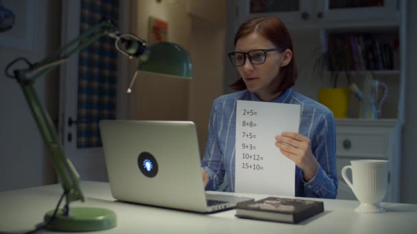 Eine junge Lehrerin in den Dreißigern mit Brille spricht und zeigt ein Blatt Papier mit einfachen mathematischen Gleichungen, das zu Hause auf den Laptop schaut. Online-Bildungsprozess. Arbeit von zu Hause aus. — Stockvideo