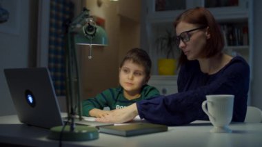 30 'lu yaşlarda genç bir anne, okullu oğluyla çevrimiçi ödev yapıyor. Gözlüklü bir kadın ve dizüstü bilgisayarı, defteri ve ders kitabı olan bir çocuk. Çevrimiçi eğitim süreci. 