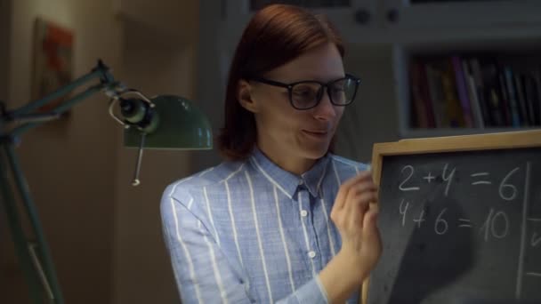 Femme des années 30 en lunettes écrivant des mathématiques sur un tableau de craie avec de la craie blanche regardant la caméra à la maison. Processus d'éducation en ligne. Webcam vue de professeur parler . — Video