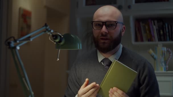 Homem de 30 anos barbudo em óculos mostrando livro de papel e explicando na câmera em casa. Processo de educação online. Webcam vista de professor falando . — Vídeo de Stock
