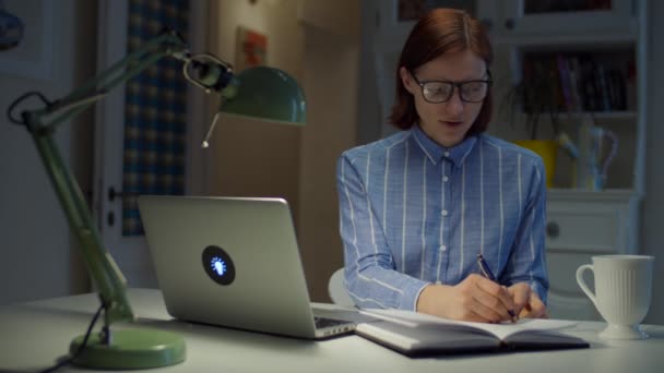 Junge Frauen in den 30er Jahren machen sich von zu Hause aus Notizen mit Stift und reden online per Laptop. Online-Bildungsprozess. Lächelnde Geschäftsfrau mit Brille zu Hause. — Stockvideo