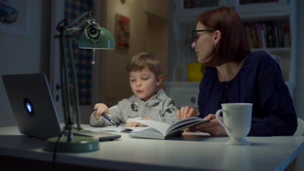 Junge Mutter in den 30er Jahren macht mit ihrem Vorschulsohn zu Hause Online-Hausaufgaben. Frau mit Brille und kleiner Junge beim Online-Lernen mit Laptop, Notizbuch und Lehrbuch. Online-Schulung. — Stockvideo