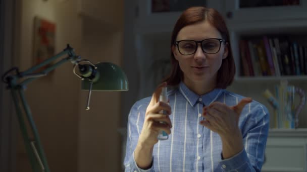 Νεαρή 30άρα γυναίκα με γυαλιά δείχνει πώς να χρησιμοποιεί απολυμαντικό χεριών και χαμογελώντας κοιτάζοντας την κάμερα. Θηλυκό απολυμαντικό στο σπίτι σε αυτο-απομόνωση από τον ιό. — Αρχείο Βίντεο