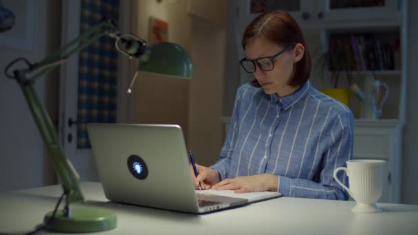 Jonge dertigers werken vanuit huis vrouw maakt notities met pen gericht op laptop scherm. Online onderwijsproces. Zakenvrouw met bril thuis. — Stockvideo