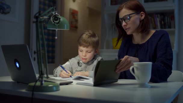 Giovane madre degli anni '30 che fa i compiti online con il figlio in età prescolare a casa. Donna in occhiali e ragazzo che studia online con laptop, notebook e libro di testo. Processo educativo online . — Video Stock