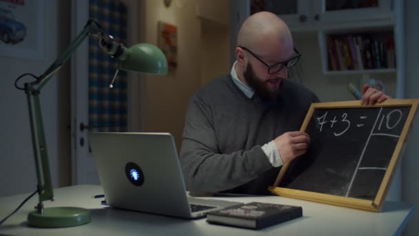 戴眼镜的30多岁的男教师，在粉笔板上写数学，白色粉笔看着家里的笔记本电脑。在线教育进程。教师谈话的侧面观点. — 图库视频影像
