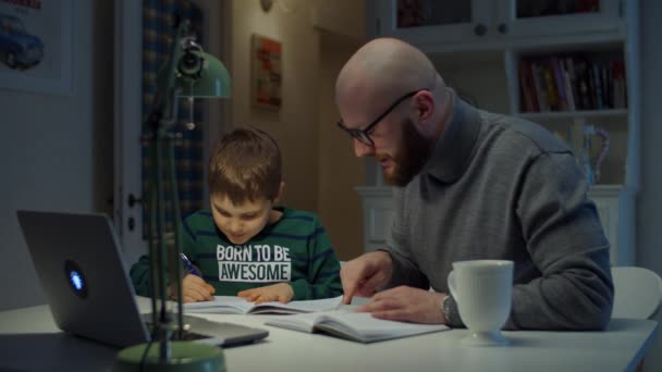 Young 30s father making online homework with schooler son at home. Bearded man in glasses and schoolboy study online with laptop, notebook and textbook. Online education process. — Stock Video