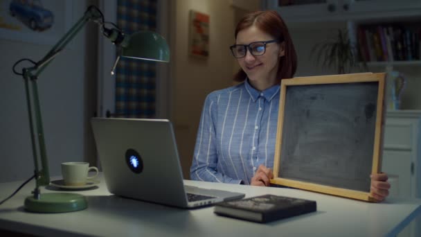 Eine junge lächelnde Frau in den Dreißigern mit Brille, die eine leere Kreidetafel in der Hand hält und zu Hause auf den Laptop schaut. Online-Bildungsprozess. Seitenansicht des Lehrergesprächs. — Stockvideo