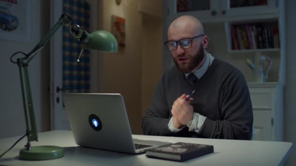 Barbudo hombre de 30 años en gafas habla emocionalmente en el ordenador portátil durante la videollamada en casa. Proceso de educación en línea. Vista lateral del profesor hablando . — Vídeo de stock