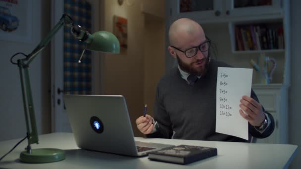 Mężczyzna po trzydziestce w okularach pokazuje kartkę papieru z prostymi równaniami matematycznymi patrzący na laptopa w domu. Proces edukacji online. Widok mówienia nauczyciela z boku. — Wideo stockowe
