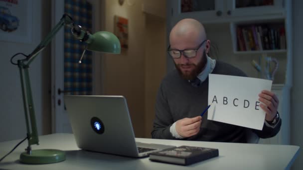 Homem de 30 anos barbudo em óculos mostrando folha de papel com letras alfabéticas olhando para laptop em casa. Processo de educação online. Visão lateral do professor falando . — Vídeo de Stock