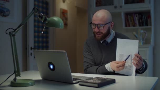 Homem de 30 anos barbudo em óculos mostrando folha de papel com gráficos para estudantes olhando para laptop em casa. Processo de educação online. Visão lateral do professor falando . — Vídeo de Stock