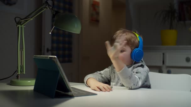 Niño preescolar en auriculares de color utilizando tableta para la educación en línea en el hogar. Niño disfruta de hacer la tarea de la escuela en línea, respuestas a la pantalla del gadget . — Vídeos de Stock