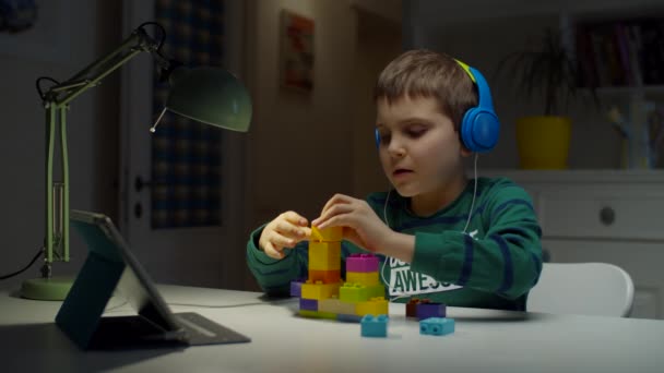 Niño colegial en auriculares de color con constructor utilizando tableta de computadora para la educación en línea en el hogar. Niño disfruta jugando con ladrillos constructores con gadget . — Vídeos de Stock