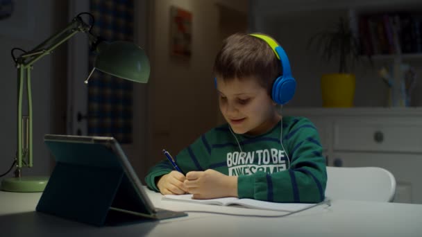 Schooler boy in color headphones using tablet computer for online education at home. Kid enjoys making school homework online, hand writing in textbook. — Stock Video