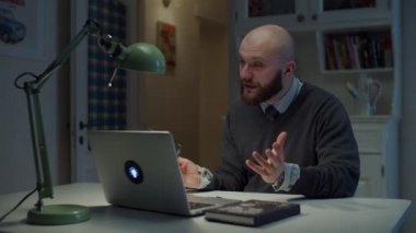 Gözlüklü 30 'lu yaşlardaki sakallı adam evdeki video görüşmesi sırasında laptopta duygusal konuşuyor. Çevrimiçi eğitim süreci. Öğretmenin konuşmasının yan görünümü.