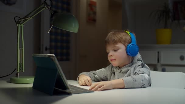 Хлопчик-дошкільнята у кольорових навушниках за допомогою планшетного комп'ютера для домашньої освіти. Малий любить робити шкільні домашні завдання онлайн, відповіді на телеекран. — стокове відео
