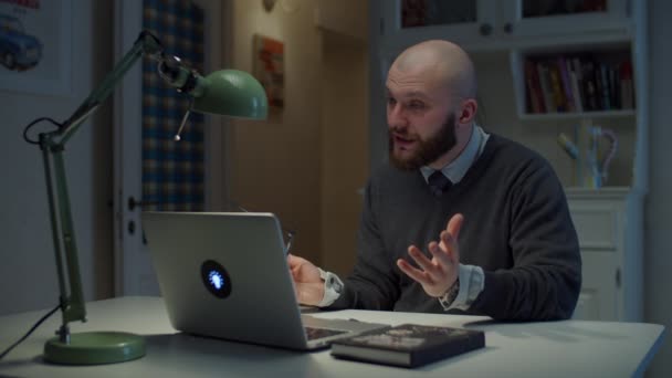 Een man van in de dertig met een bril praat emotioneel op een laptop tijdens een videogesprek thuis. Online onderwijsproces. Zijaanzicht van de leraar praten. — Stockvideo