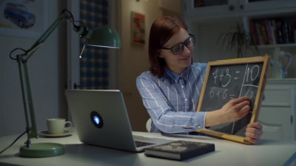 30-talets kvinnliga lärare i glasögon skriver enkla matematiska ekvationer på tavlan med vit krita tittar på laptop hemma. Utbildningsprocess på nätet. Sidovy av lärarprat. — Stockvideo