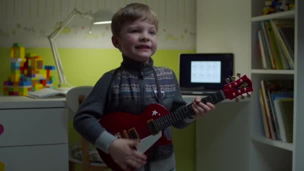 Niño rubio jugando en la guitarra de juguete y cantando mirando a la cámara con el ordenador portátil detrás. Niño tocando música en casa en cámara lenta . — Vídeo de stock