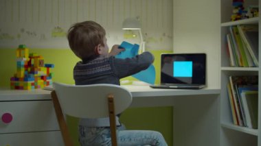 Anaokulu öğrencisi çocuk online resim ödevi yapıyor. Çocuk makasıyla mavi kare şeklini kesiyor. Evdeki çocuk odasındaki masada oturuyor..