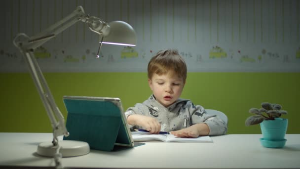 Předškolák dělá online domácí úkoly s tabletovým počítačem. Dítě sedící za stolem a studující online v dětském pokoji doma. Chlapecký rukopis v učebnici — Stock video