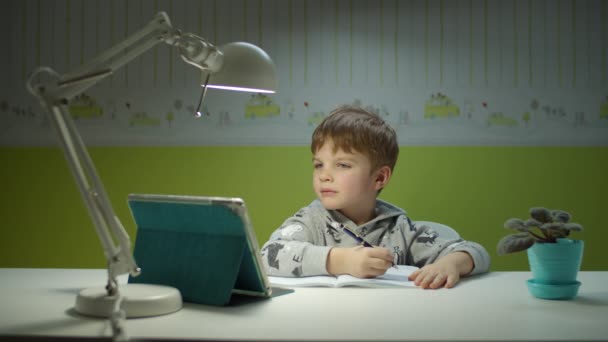 Niño preescolar haciendo tarea en línea con la computadora de la tableta. Niño sentado en el escritorio y estudiando en línea en la habitación de los niños en casa. Chico caligrafía en libro — Vídeos de Stock