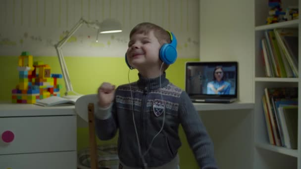 Niño preescolar en auriculares de color bailando en casa con lección en línea con la maestra en el ordenador portátil detrás. E-estudiar feliz niño bailando en la habitación de los niños . — Vídeo de stock