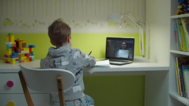 Anaokulu çocuğu dizüstü bilgisayar ekranında bayan öğretmenle online okul ödevi yapıyor. Çocuk kitaplarını raftan alıyor, masada oturuyor ve evdeki çocuk odasında internetten ders çalışıyor.. 